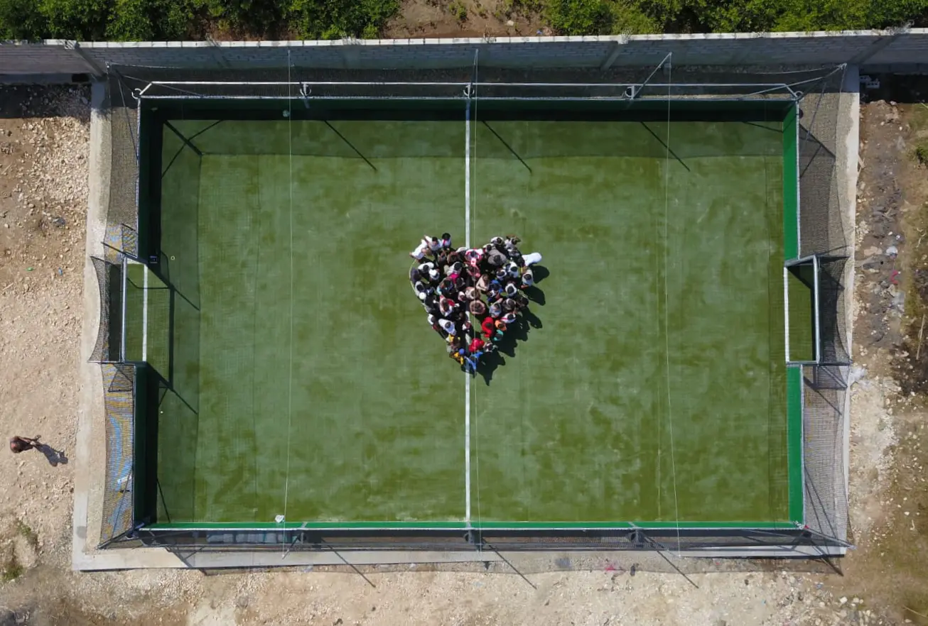Aerial view of a green mini-pitch in Haiti with children standing in a heart shape at the centre, bare ground on all sides.